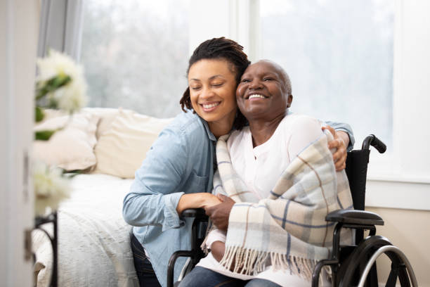 Young woman hugs senior woman in wheelchair A daughter puts her arms around a smiling mother wrapped up in a blanket, sitting in a wheelchair. healthcare worker stock pictures, royalty-free photos & images