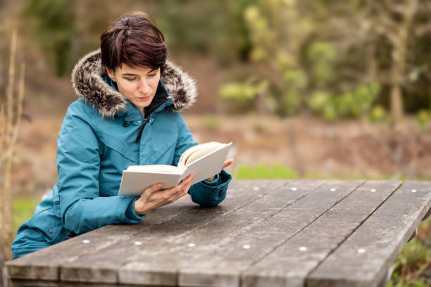 Young woman in blue raincoat reading a book outdoors sitting on a bench at a picnic table for hikers outdoors in nature stock photo