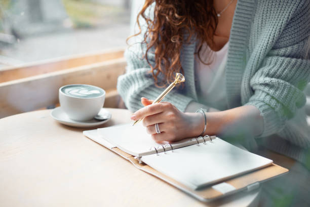 Young woman in blue warm sweater sitting near the big window of coffee shop and writing notes stock photo