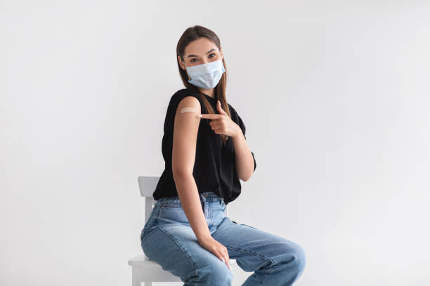 Young woman in face mask pointing at adhesive bandage on arm, getting vaccinated against covid-19 on light background stock photo