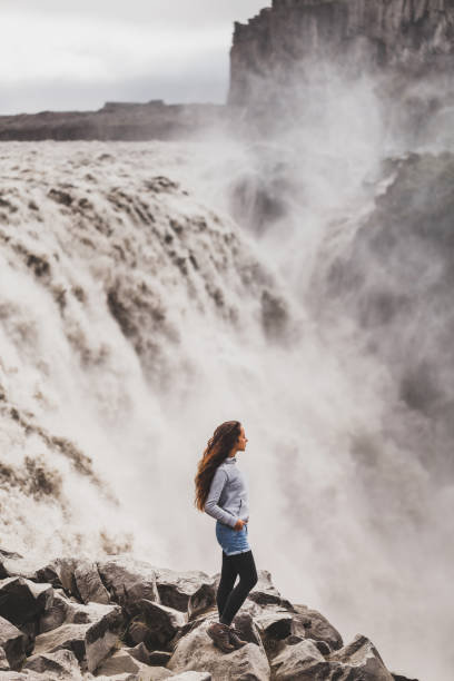Young woman in jeans shorts and gray hoodie with dramatic view of famous Iceland waterfall Dettifoss. Breathtaking landscape, stream of water, most powerful waterfall in Europe. Small person, big waterfall. Young woman in jeans shorts and gray hoodie with dramatic view of famous Iceland waterfall Dettifoss. Breathtaking landscape, stream of water, most powerful waterfall in Europe. Small person, big waterfall. iceland dettifoss stock pictures, royalty-free photos & images