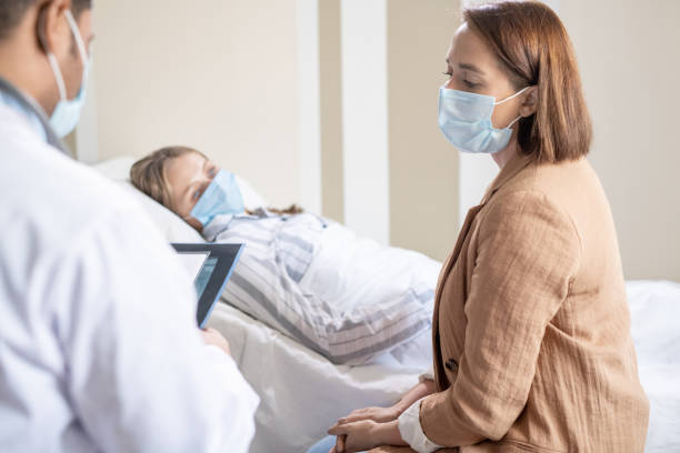 Young woman in protective mask consulting with doctor about her sick friend Young brunette woman in protective mask consulting with doctor about her sick friend lying in bed near by in contemporary covid hospital visit stock pictures, royalty-free photos & images