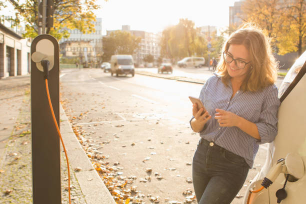 ung kvinna stående nära elbilen och hålla smartphone - electric car woman bildbanksfoton och bilder