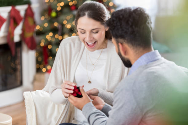 Young woman is surprised as her boyfriend proposes to her Excited young woman accepts her boyfriend's marriage proposal on Christmas Eve. He is kneeling while giving her the ring. fiancé stock pictures, royalty-free photos & images