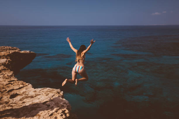 Young woman jumping off cliff and diving into blue sea Young active woman jumping off cliff and falling into ocean on tropical island summer holidays cliff jumping stock pictures, royalty-free photos & images