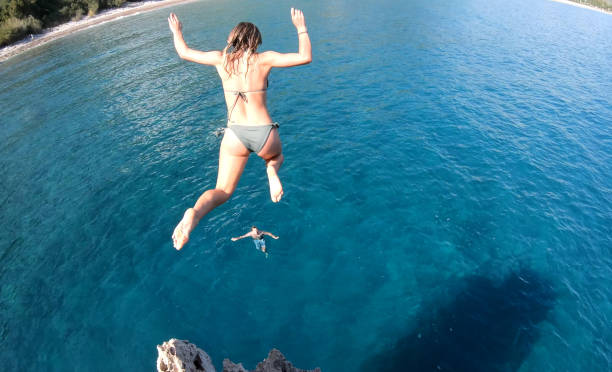 Young woman jumps off high cliff into the sea Young man swims in shallows below cliff jumping stock pictures, royalty-free photos & images