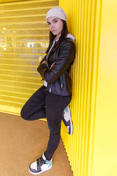 A young woman leaning against a yellow wall with her arms crossed and looking at the camera stock photo