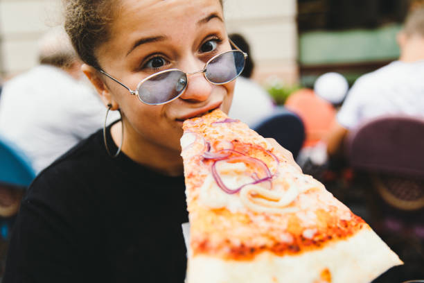 Young woman likes her pizza so much! Young funny woman with sunglasses and curly hair eating a big piece of tasty pizza outdoors at summer terrace of cafe lviv photos stock pictures, royalty-free photos & images