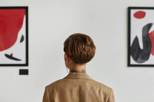 Young Woman Looking at Modern Art Back View Graphic back view portrait of tattooed young woman looking at paintings and listening to audio guide at modern art gallery exhibition, copy space exhibition photos stock pictures, royalty-free photos & images