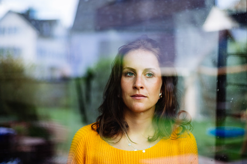 Young woman looks thoughtfully and sadly through the window into the garden with children's toys