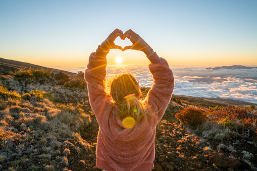 Young woman on a volcano looking at sun setting over the cloudscape and making a heart shape finger frame