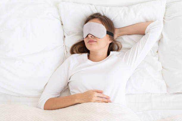 Young woman lying on bed with spread elbows, having eyemask on to dream in daylight, sleeping in calm atmosphere, relaxing Young woman lying on bed with spread elbows, having eyemask on to dream in daylight, sleeping in calm atmosphere, relaxing eye mask stock pictures, royalty-free photos & images