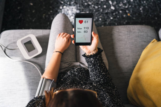 Young woman measures blood pressure sitting on sofa at home with smartphone connected to device - Concept of health, well-being and love for oneself - Millennial in a moment of private life stock photo