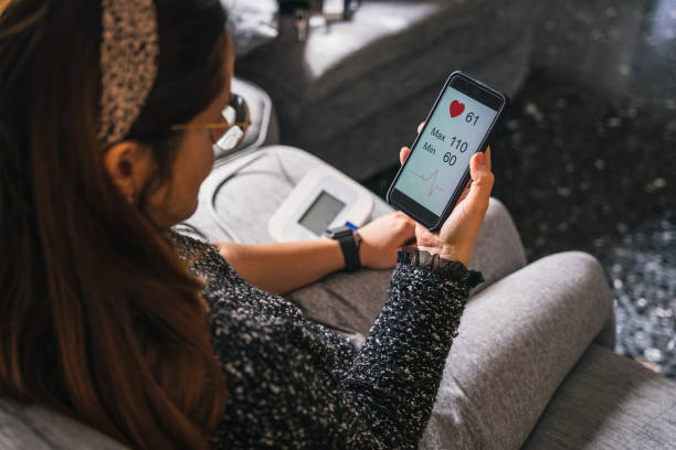 Young woman measures blood pressure sitting on sofa at home with smartphone connected to device - Concept of health, well-being and love for oneself - Millennial in a moment of private life stock photo