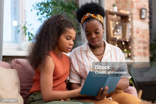 Young woman of African ethnicity and her cute teenage daughter reading book