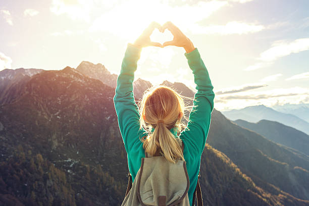 Young woman hiking on the mountain top makes a heart shape finger frame above her head to the beautiful landscape.