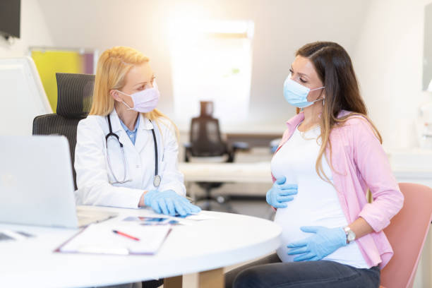Young woman on pregnancy exam during Covid 19. Young pregnant woman on a visit to doctor. Wearing protective masks and gloves during corona virus epidemic. Doctor listening to baby with stethoscope. obstetrician photos stock pictures, royalty-free photos & images