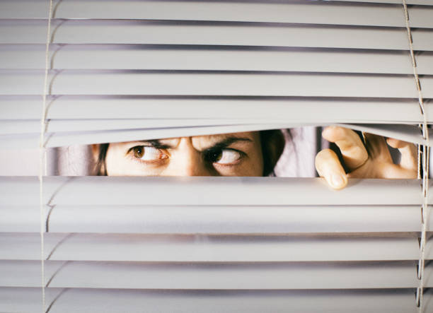 Young woman peering anxiously through blinds stock photo