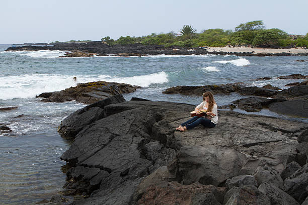 Young Woman Playing Guitar by the ocean in Hawaii "Teenage girl with long hair, sitting on lava rock next to the ocean, playing the guitar." neicebird stock pictures, royalty-free photos & images