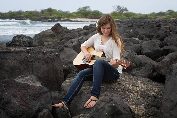 Young Woman Playing Guitar Teen girl sitting on lava rocks playing the guitar at the seashore in Hawaii. neicebird stock pictures, royalty-free photos & images