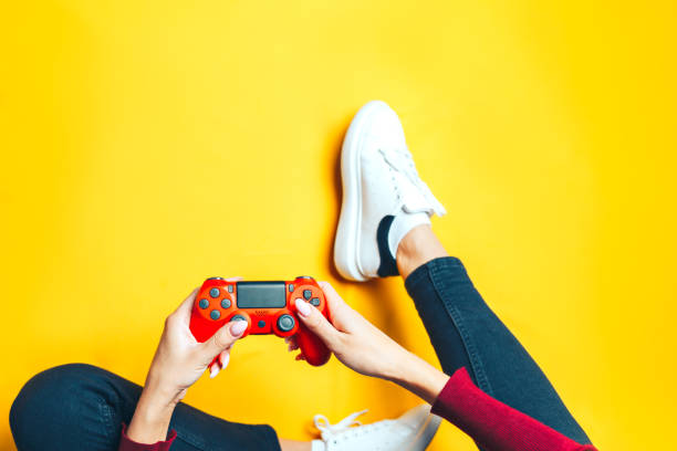 Young woman playing with two gamepads on yellow. Young thin woman playing with red gamepad, sitting on yellow background. flat lay. gambling stock pictures, royalty-free photos & images