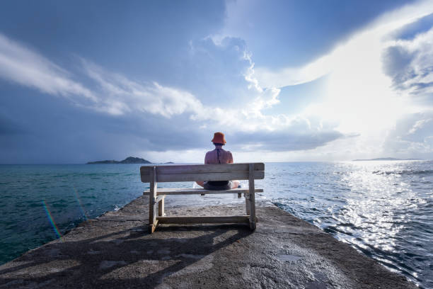 Young woman reads a book on a bench at a harbor near the sea with dramatic sky in the background stock photo