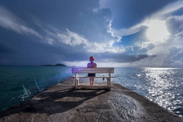 Young woman reads a book on a bench at a harbor near the sea with dramatic sky in the backgroung stock photo