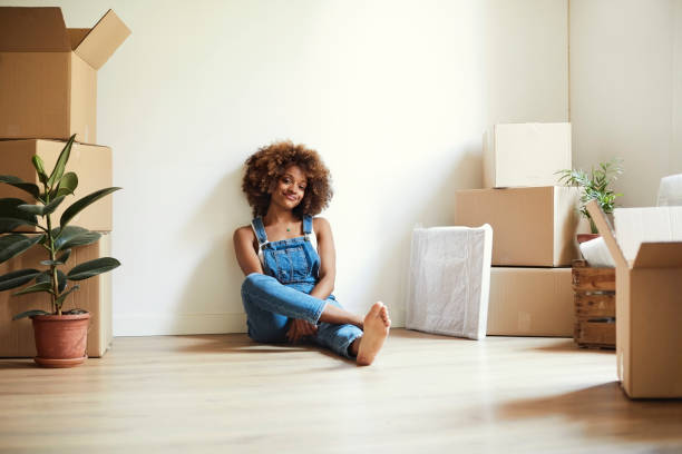 Young woman relaxing amidst moving boxes in house Full length of young woman sitting on floor in new house. Portrait of happy female is relaxing amidst moving boxes. She is wearing bib overalls. one young woman only stock pictures, royalty-free photos & images