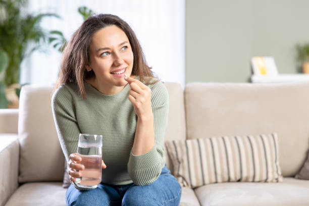 Young woman relieving disease symptoms taking a pill Smiling young woman relieving disease symptoms taking a painkiller pill sitting on sofa of in living room allergy medicine stock pictures, royalty-free photos & images