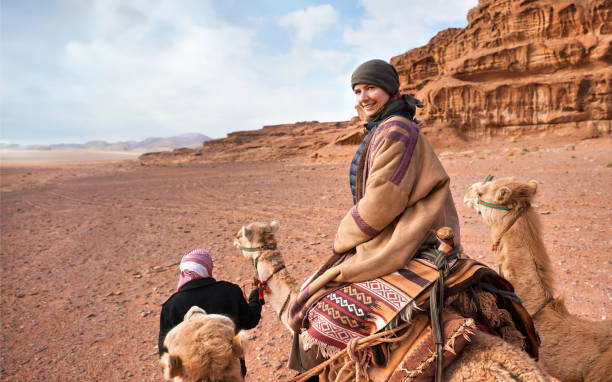 Young woman riding camel in Wadi Rum desert, looking back over her shoulder, smiling. It's quite cold so she is wearing traditional Bedouin coat - bisht - and head scarf, mountains far background Young woman riding camel in Wadi Rum desert, looking back over her shoulder, smiling. It's quite cold so she is wearing traditional Bedouin coat - bisht - and head scarf, mountains far background hot arab woman stock pictures, royalty-free photos & images