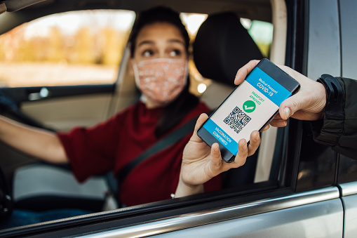 Young woman in a car showing a smartphone with a digital international certificate of Covid-19 Vaccination to an unrecognizable custom official.