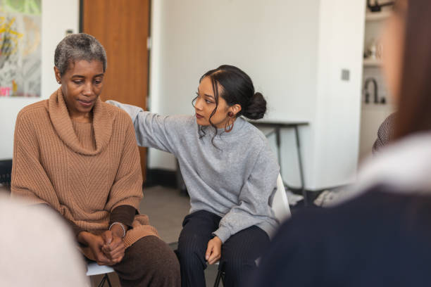 Young woman shows support in therapy session A multi-ethnic group of adults are attending a group therapy session. The attendees are seated in a circle. A senior black woman is sharing her struggles with the group. A mixed-race young woman rests her arm on the woman's back, expressing comfort and support. grief stock pictures, royalty-free photos & images