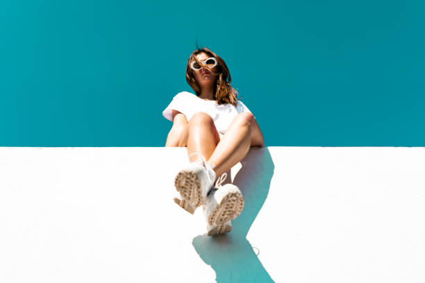 Young Woman Sitting on Concrete Wall Young woman wearing large white sunglasses sitting on modern concrete wall. Simplicity Modern Urban Style. Shot from below against the sky. Urban Lifestyle Youth Portrait. t shirt photos stock pictures, royalty-free photos & images