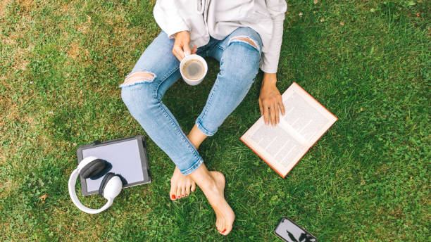 Young woman sitting on the grass drinking coffee and reading a book enjoys outdoor recreation. stock photo