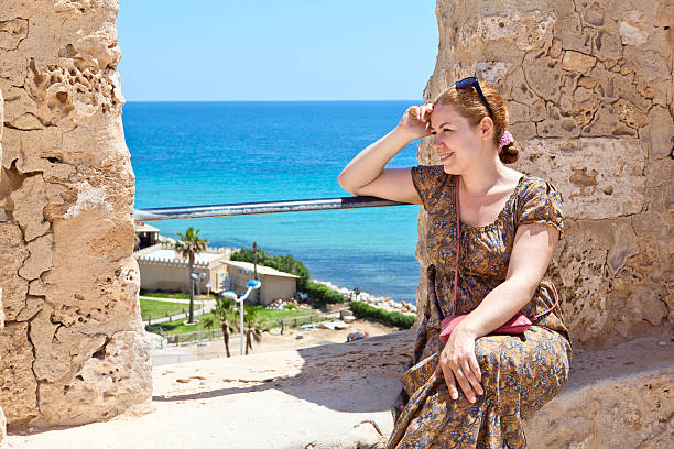 Young woman sitting on wall of ancient fortress Young woman with sunglasses sitting on wall of ancient fortress and smiling tunisia woman stock pictures, royalty-free photos & images