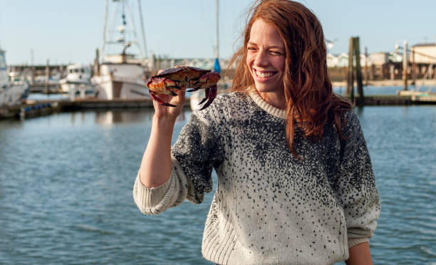 Young woman smiles as she holds up crab she caught She stands on a pier, harbour in background crabbing stock pictures, royalty-free photos & images