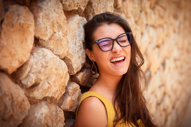 young woman standing in front of a stone wall. wearing glasses making a funny face at the camera stock photo
