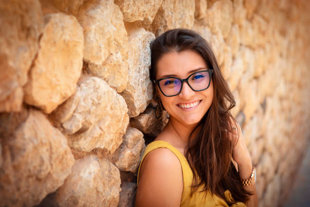 young woman standing in front of a stone wall. wearing glasses making a funny face at the camera stock photo
