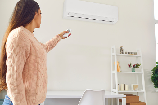 People make life comfortable. Young woman in sweater standing in living-room and switching on air conditioner with remote control to regulate temperature at home and enjoy fresh warm or cool airflow