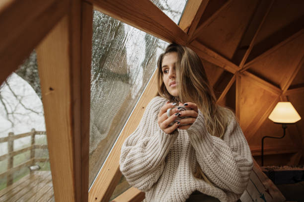 jonge vrouw die zich met een warme drank door een houten koepelvenster bevindt - rain woman sun stockfoto's en -beelden
