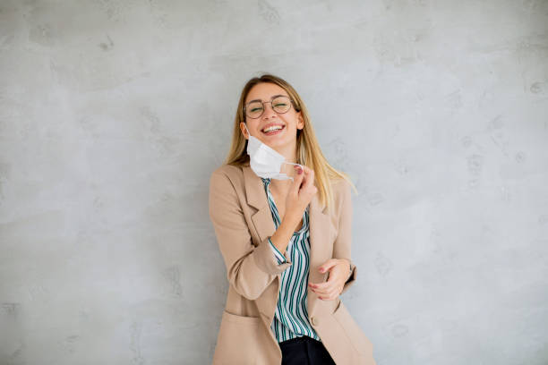 Young woman standning by the grey wall and taking off a respiratory mask from coronavirus disease stock photo
