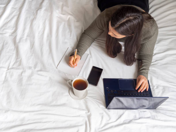 Young woman studying and working from bed with laptop, cellphone and a coffee. stock photo