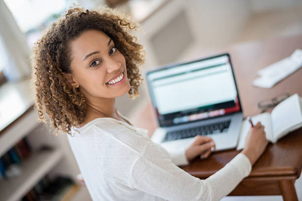 Young woman studying at home Young woman studying at home on a laptop computer and looking at the camera smiling online colleges stock pictures, royalty-free photos & images