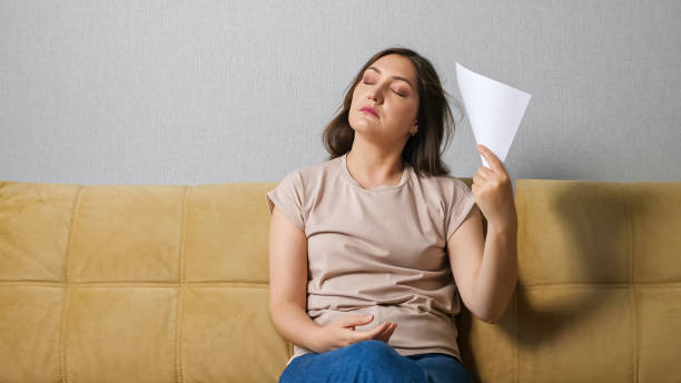 Young woman suffering from heat fan herself with a sheet of paper while sitting on the couch stock photo
