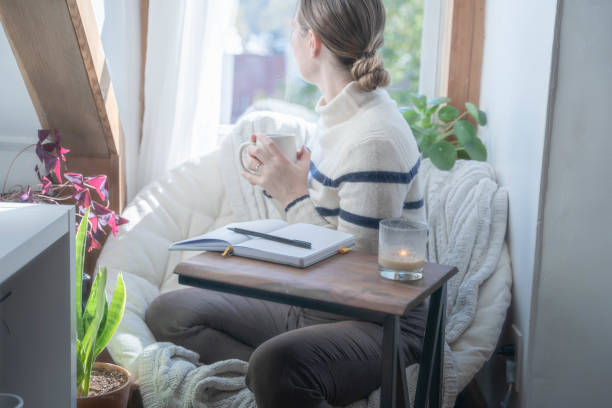 Young woman taking a mental health break to write in her journal stares out the window with coffee in hand stock photo