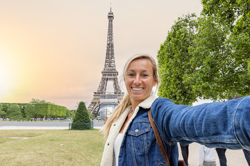 She holds her mobile phone and smiles at the camera, portrait of blond hair woman at the international landmark in Paris, France