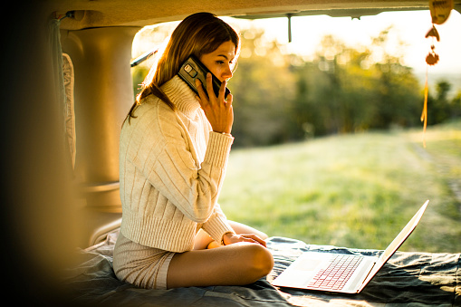 Young Woman Talking on Mobile Phone and Using Laptop on Camping Trip.