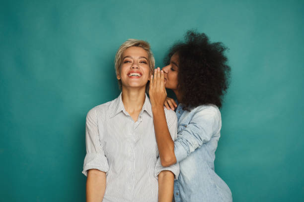 Young woman telling her friend some secrets Young woman telling her girlfriend some secret. Two women gossiping. Excited emotional girl whispering to her friend ear, turquoise studio background girlfriend stock pictures, royalty-free photos & images