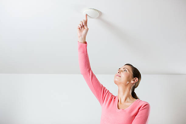 young woman testing smoke alarm on ceiling - rookmelder stockfoto's en -beelden