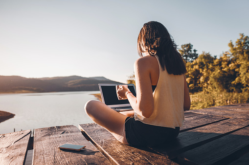 Woman working on laptop in the nature, she sitting on wooden pier around beautiful mountain lake and using laptop in the morning